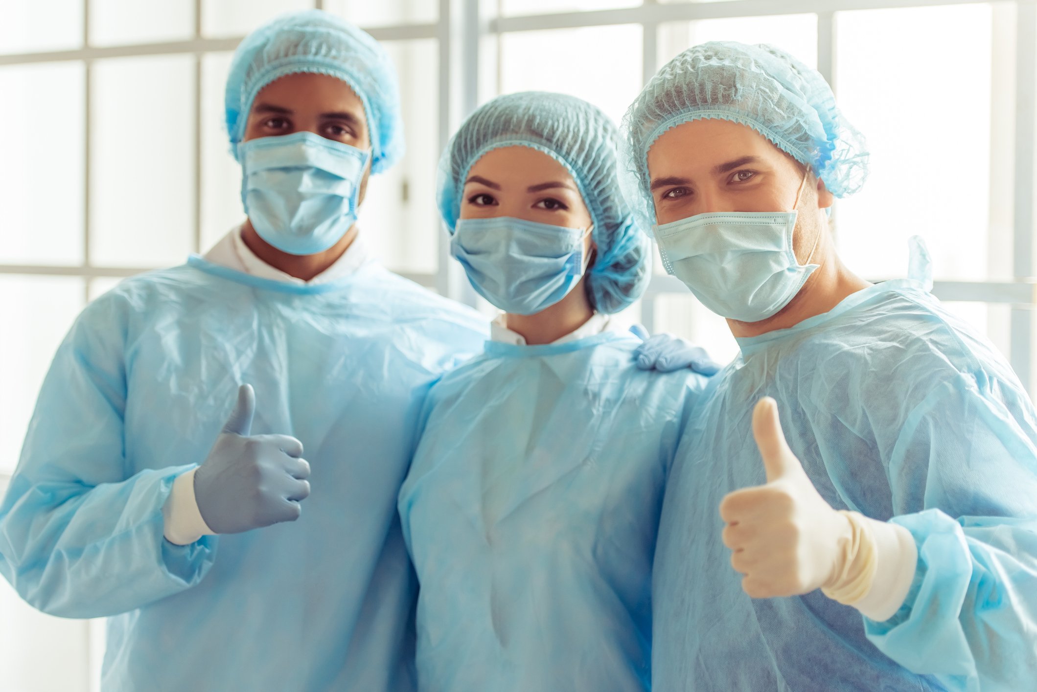 Three healthcare workers in scrubs, two giving a thumbs-up.