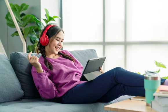 Teen girl wearing headphones watching  a program on her tablet while propped up on couch. 