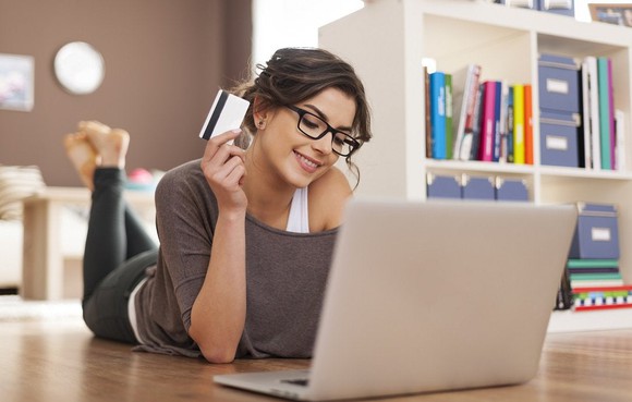 A young woman in glasses lays on the floor holding a credit card and using a laptop.