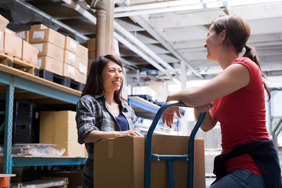 Two women chatting while loading a box onto a trolley in a warehouse.