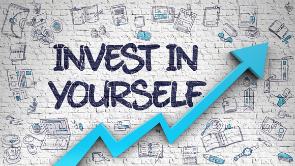 The words "Invest in Yourself" written on a brick wall with an upward trending blue arrow underneath. 