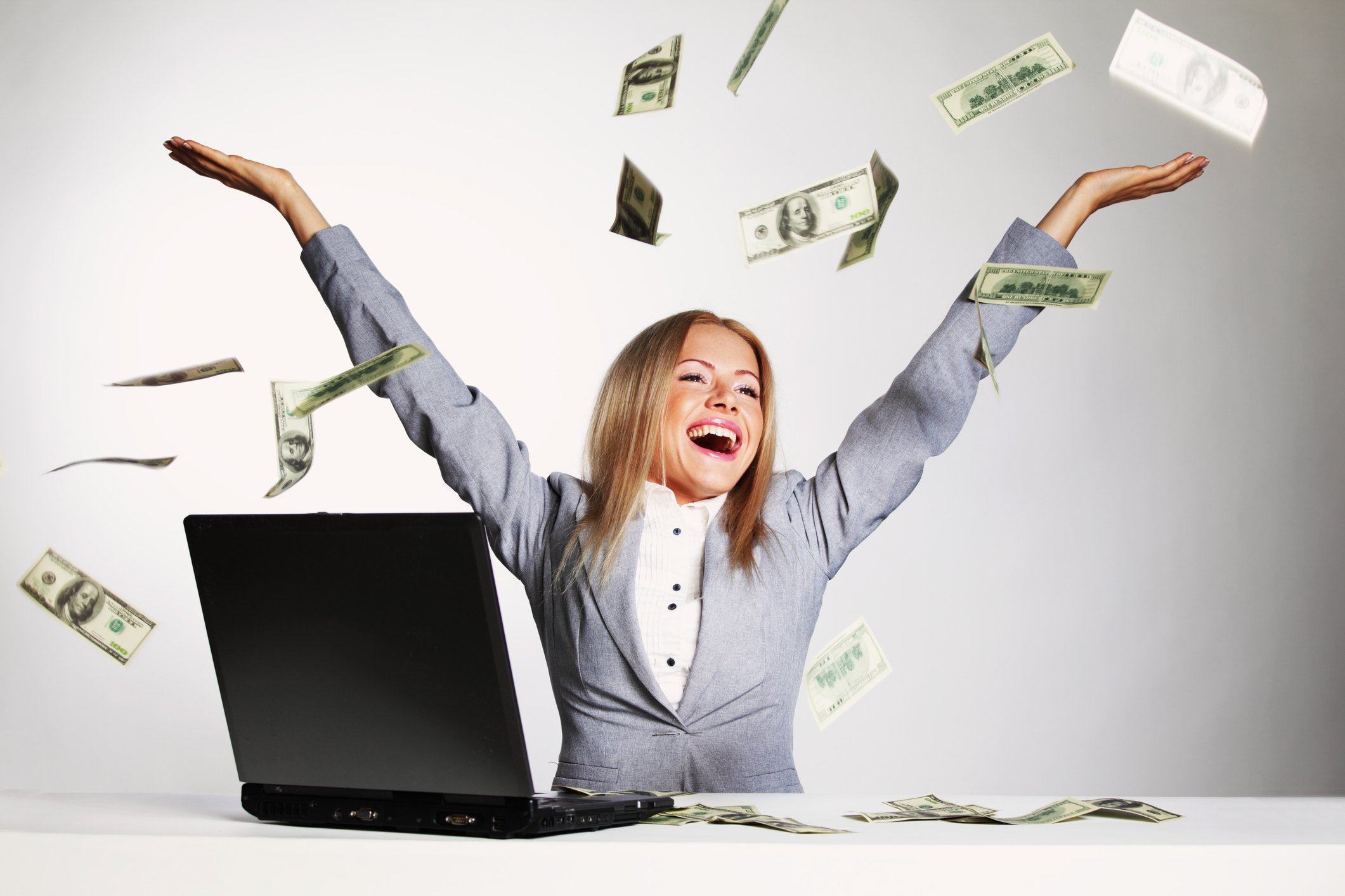 A young businessperson sits at a laptop, throwing hundred-dollar bills in the air with a big smile.