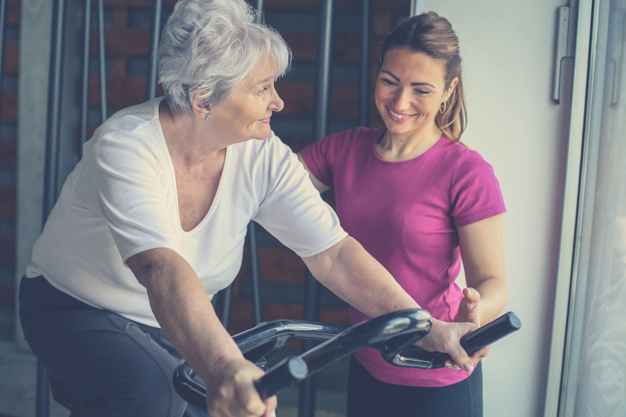 Old lady on exercise bike while younger lady smiles and looks on