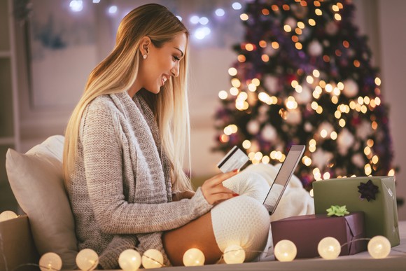 A woman, sitting in front of a Christmas tree, smiles as she shops online.