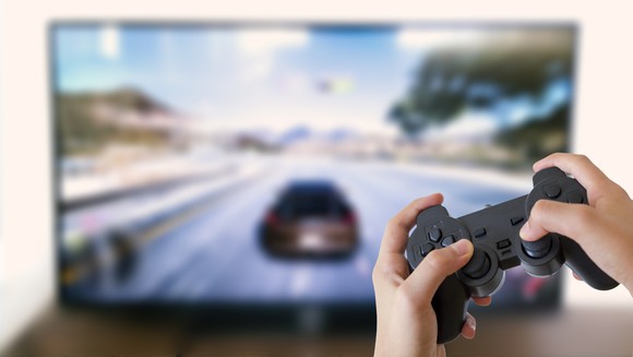 A person's hands holding a video game controller with the game displayed on a TV in the background.