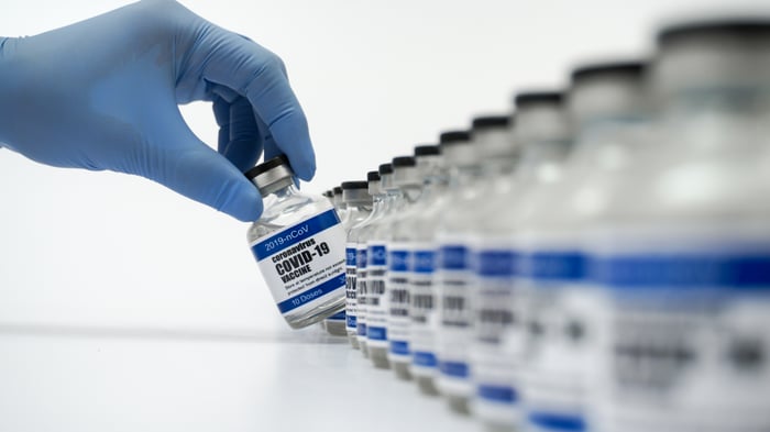 A gloved hand selects one of a row of vials labeled Coronavirus COVID-19 Vaccine.