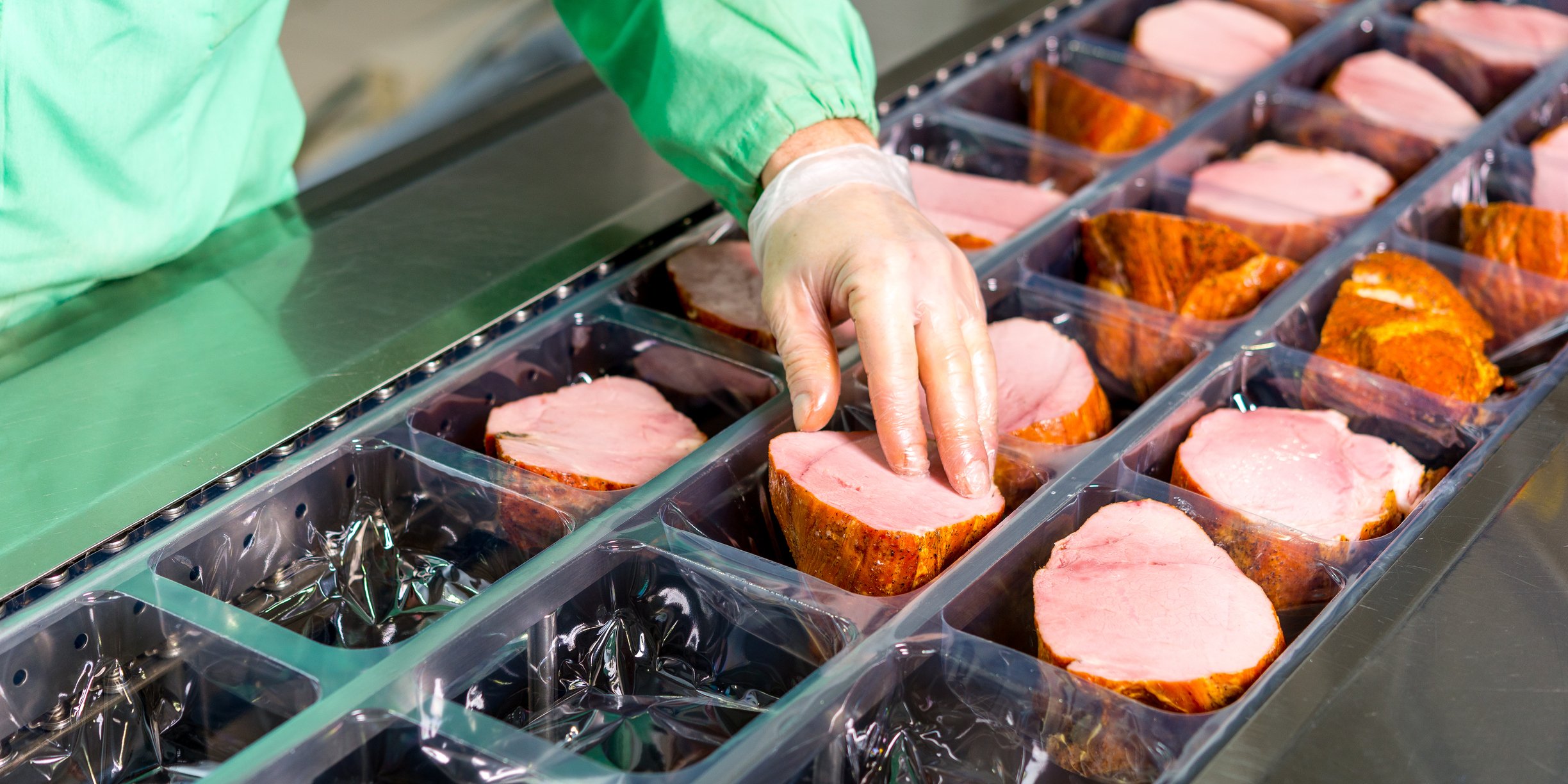 A worker wearing latex gloves and protective clothing inspecting hams before packaging.