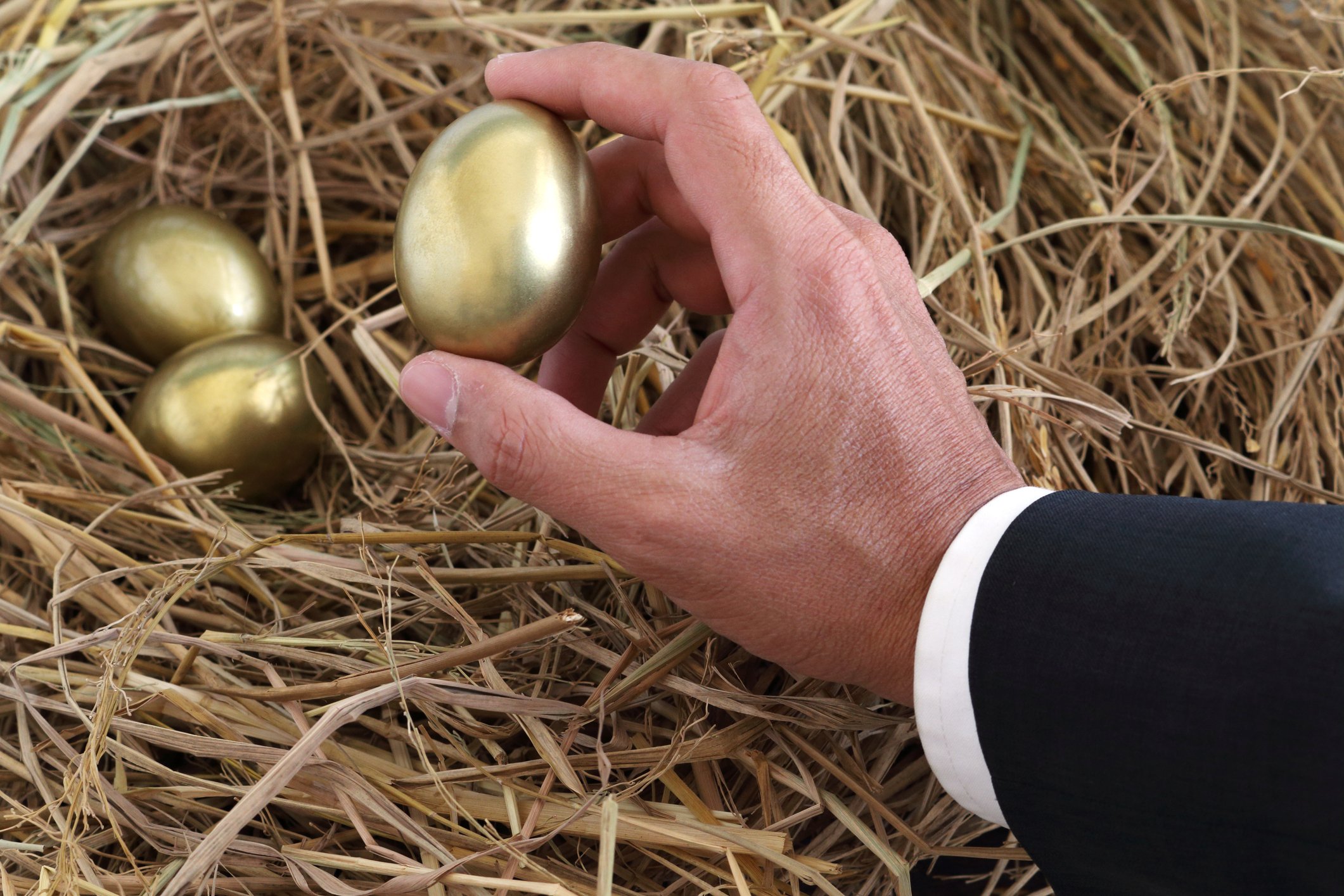 Close-up photo of a businessman's hand picking up a golden egg from a nest.