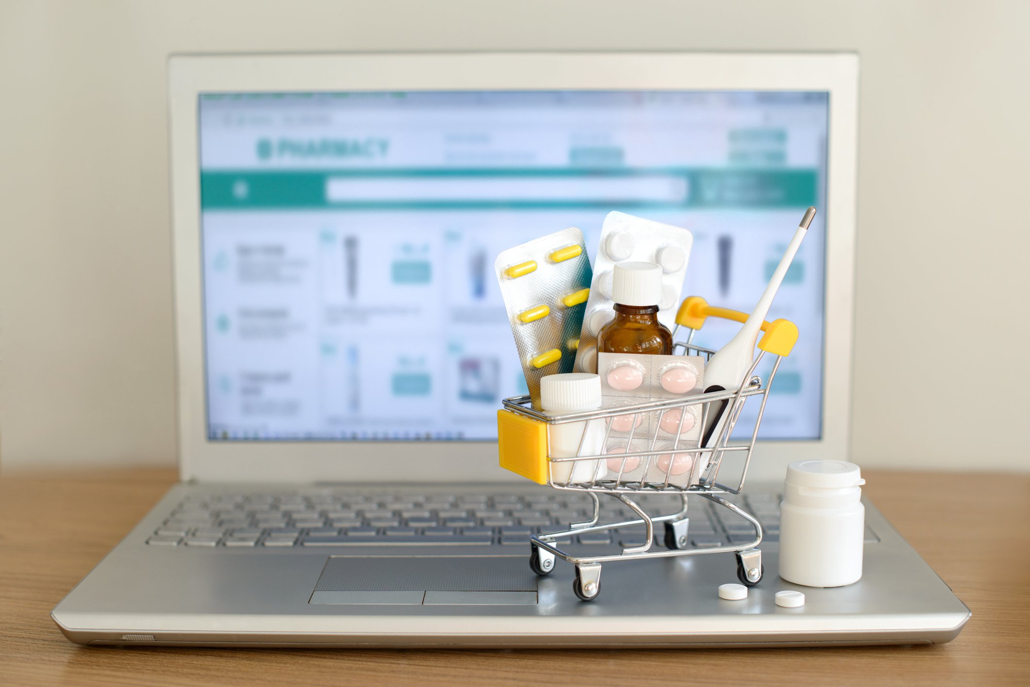 A miniature shopping cart filled with prescription drugs sits on atop a laptop displaying an online pharmacy 