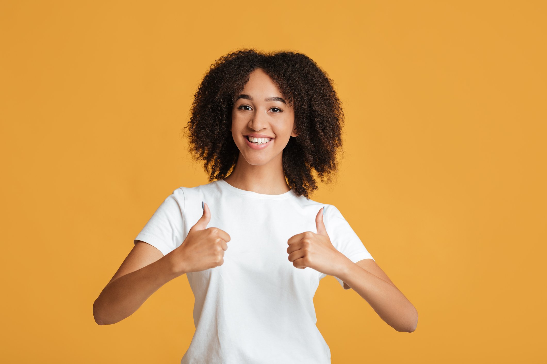 A woman holdings two thumbs up and smiling in front of a yellow background. 