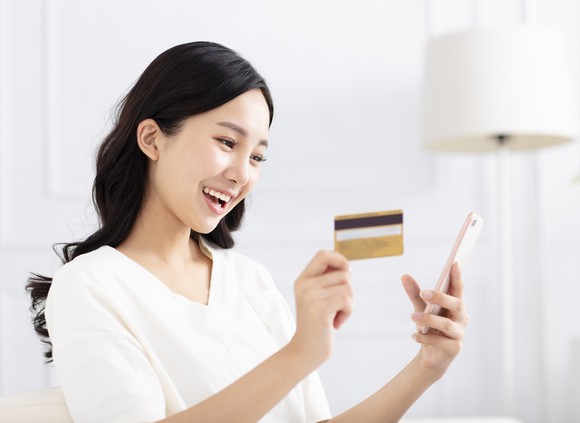 A young Asian woman smiles at her smartphone, holding a credit card in her other hand.