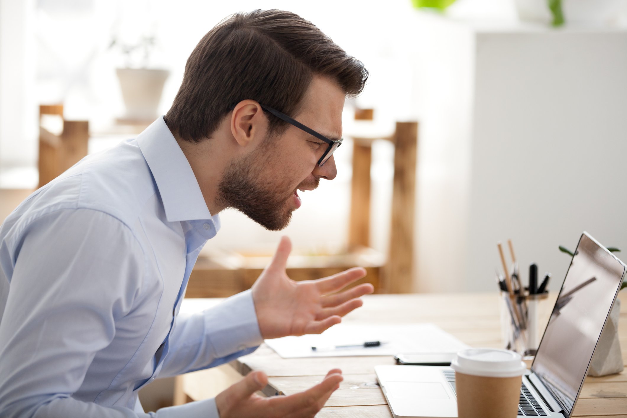 Man in dress shirt looking frustrated and angry at a laptop on a table with papers on it.