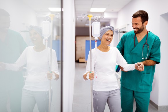 A cancer patient walking with an intravenous drip on a pole, being supported by a clinician.