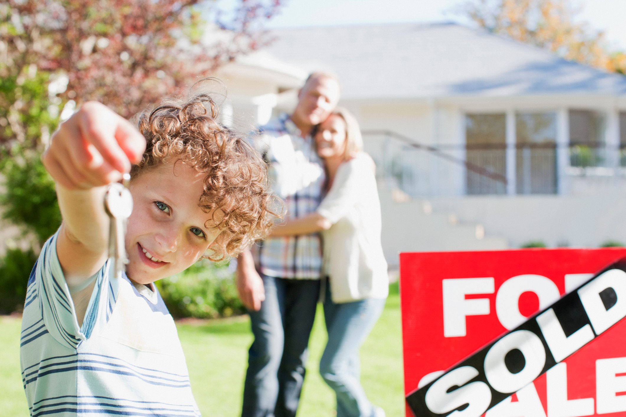 Couple standing near house sold sign, with child holding house keys nearby. 