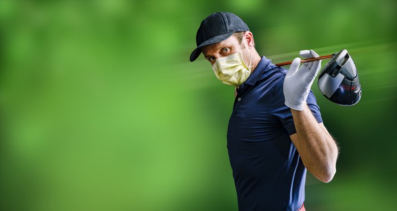 A male golfer  stretches with his driver  behind his head and wears a face mask. 