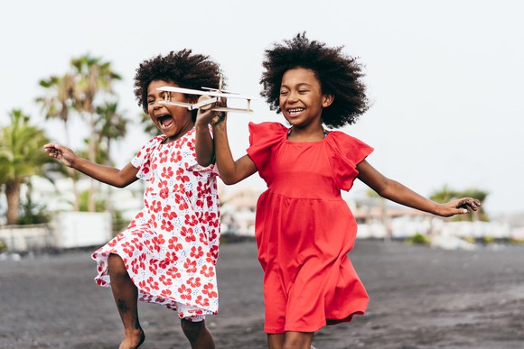 Twin African girls run with glee while holding a wooden airplane.