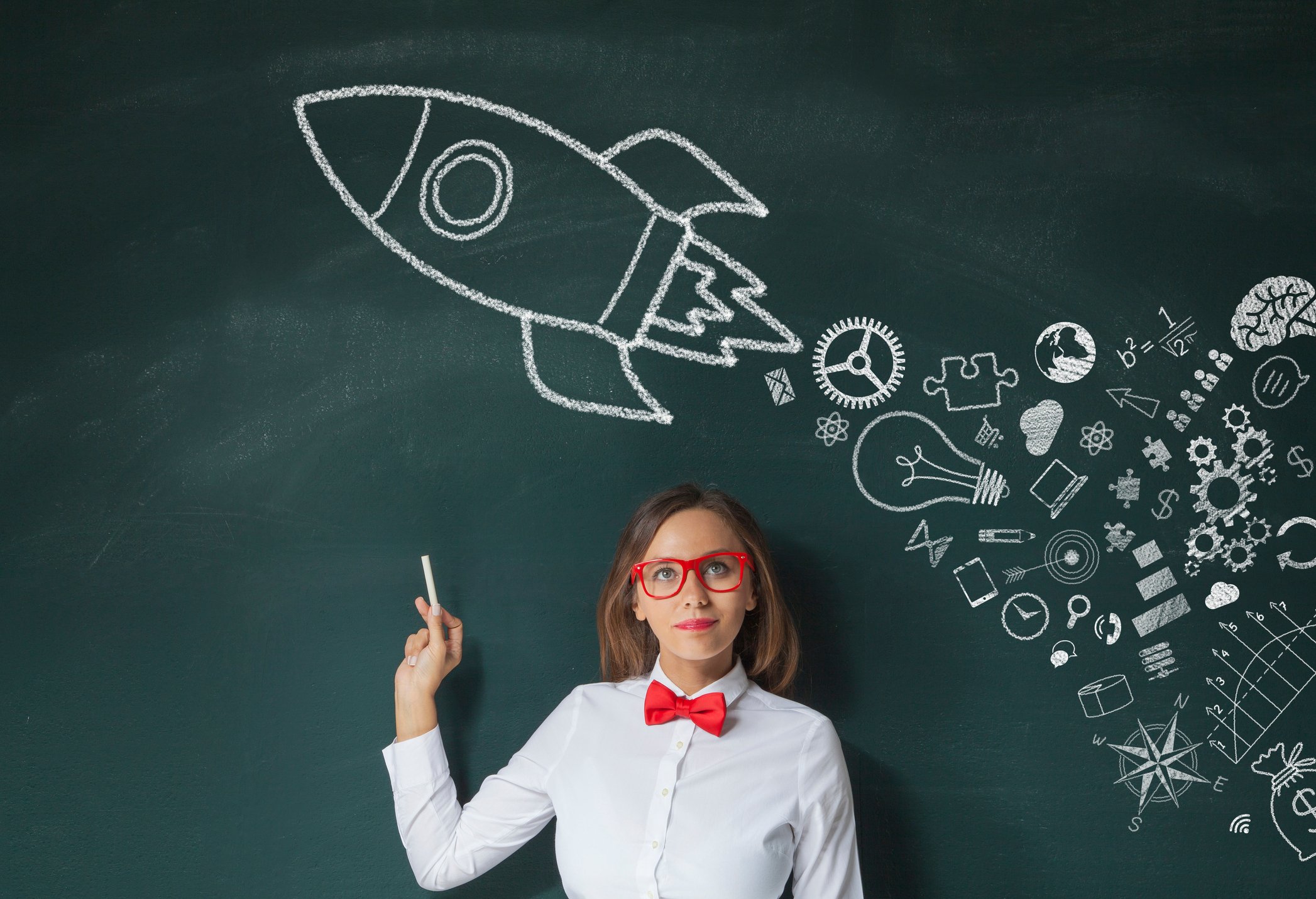 A teacher holds chalk in front of a chalkboard where a drawing of a rocket leaves drawings of various innovations in its wake instead of a smoke trail.
