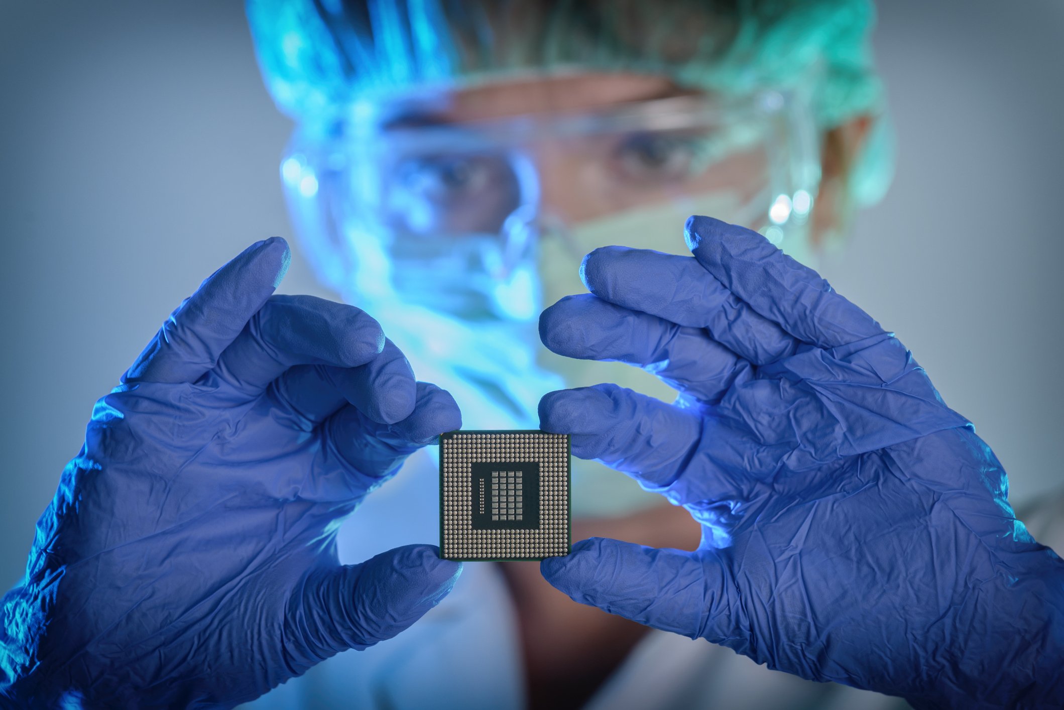 A technician in clean factory gear holds a square semiconductor chip in their hands. 