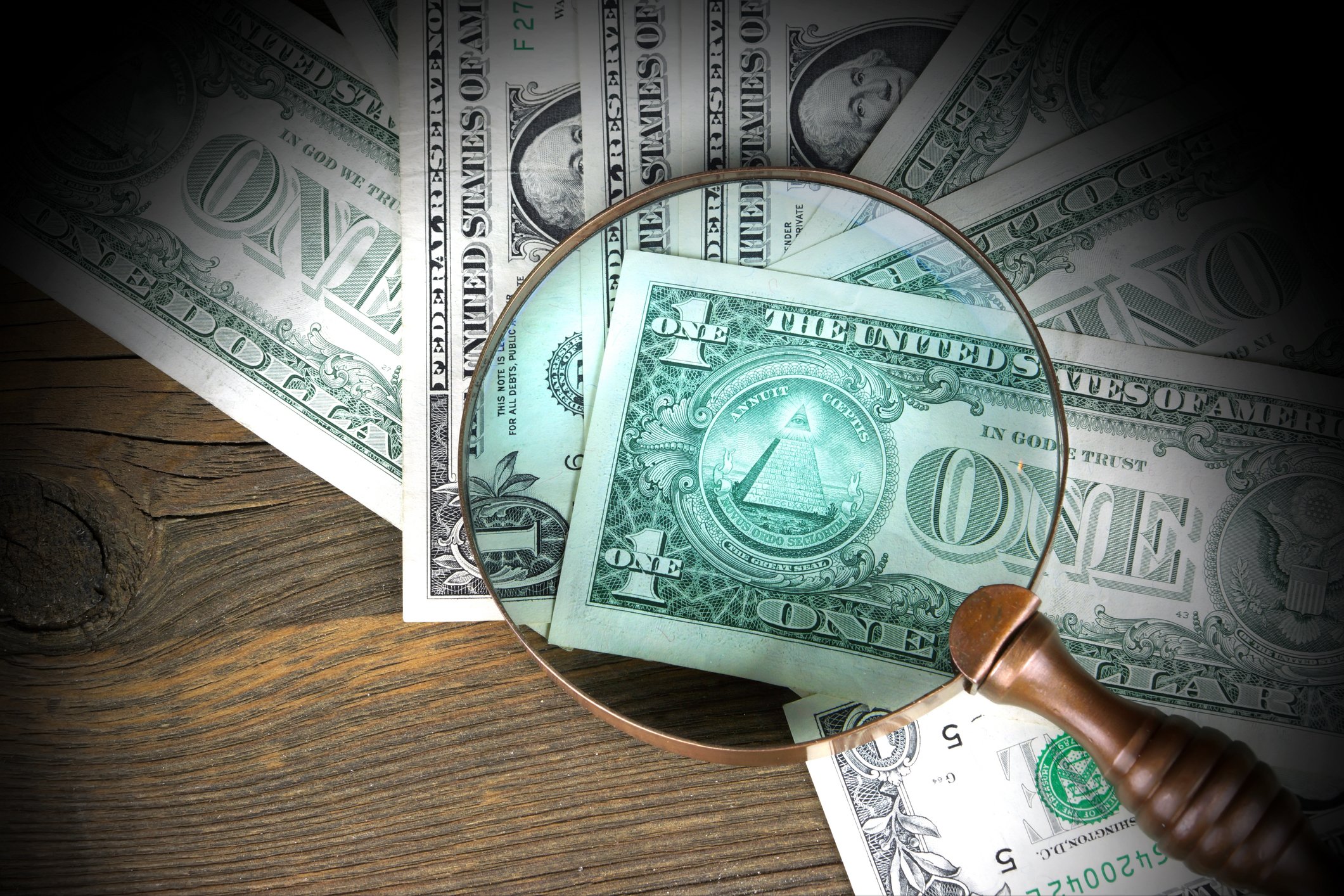 A magnifying glass highlights the eye of providence pyramid on one of several spread-out dollar bills. 