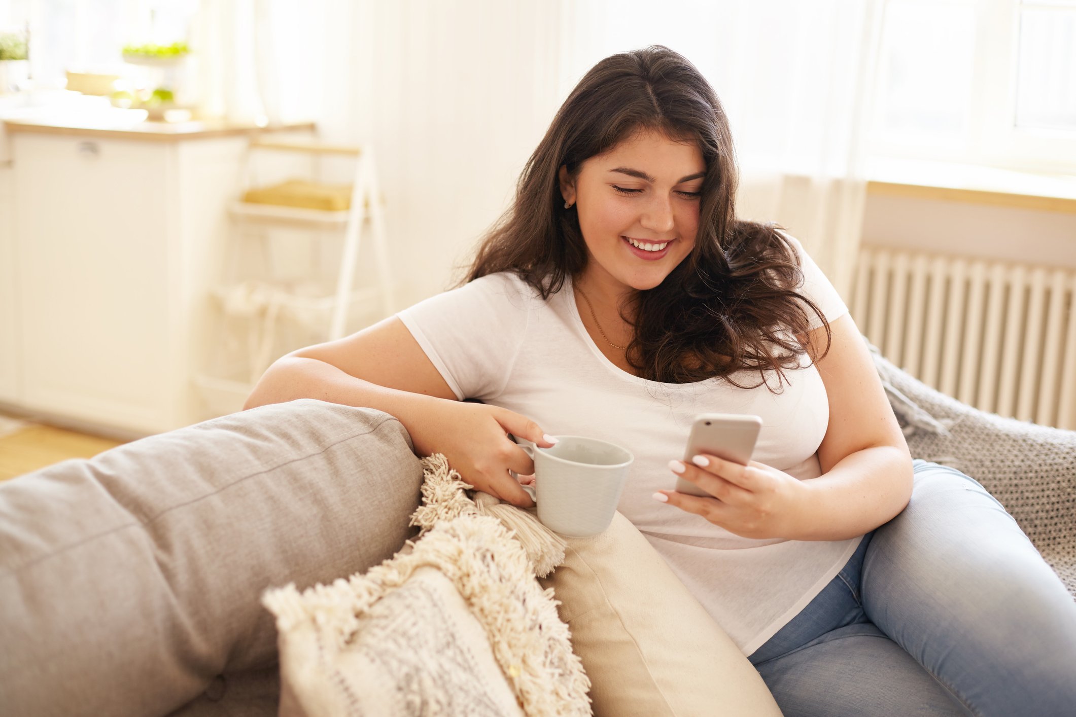 Young woman on couch staring at her smartphone while holding a cup of coffee.