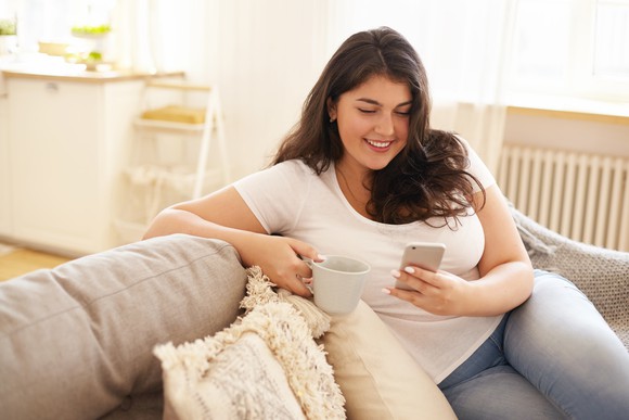 Young woman on couch staring at her smartphone while holding a cup of coffee.
