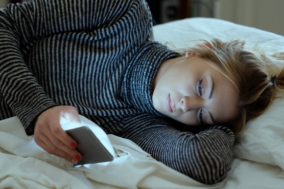 A teenage girl staring at her smartphone as she lays in bed. 