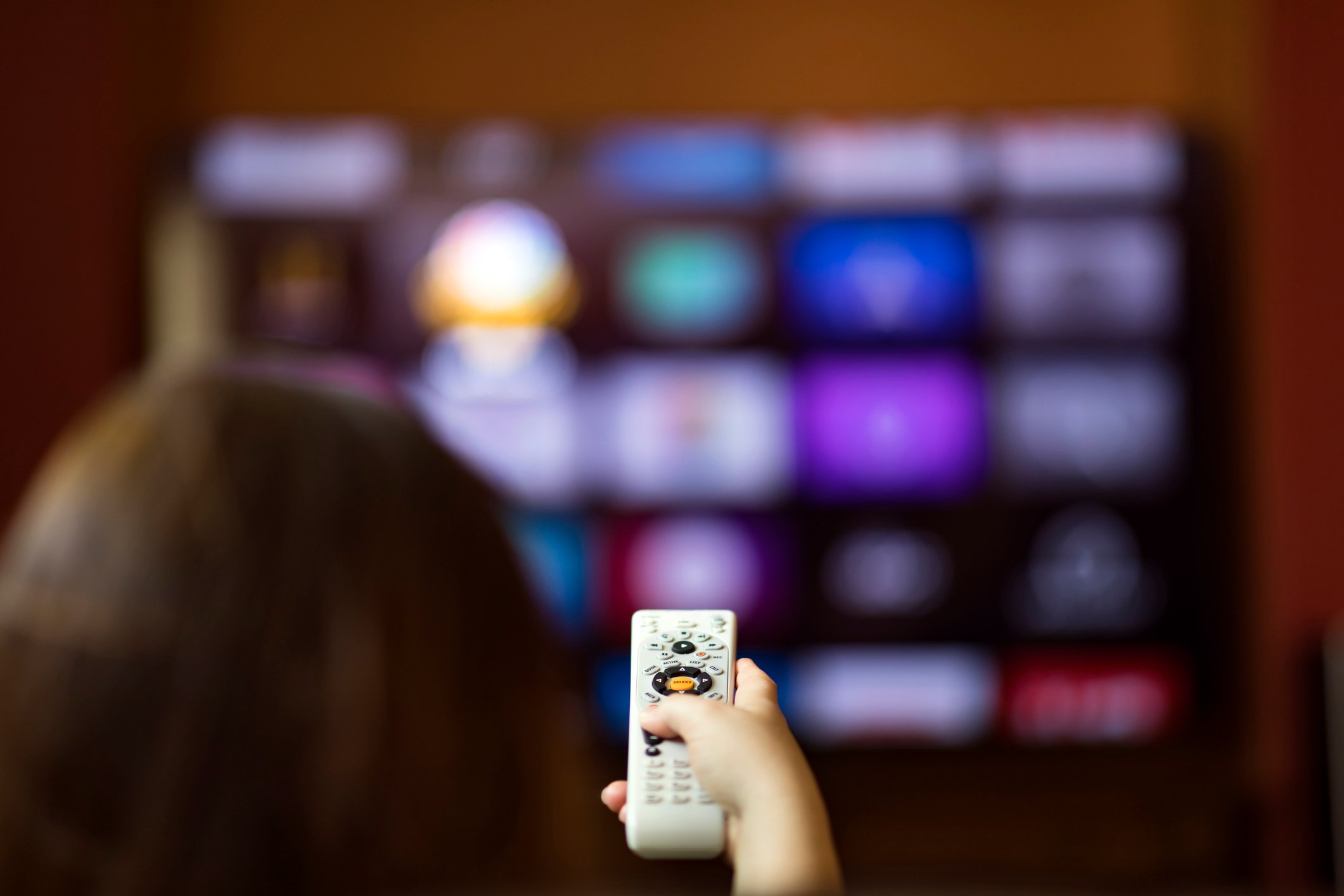 A woman in the foreground points a remote control at a TV in the background.
