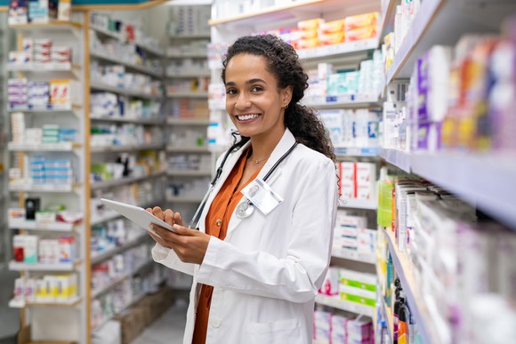 A smiling pharmacist in rows of shelving containing prescriptions.