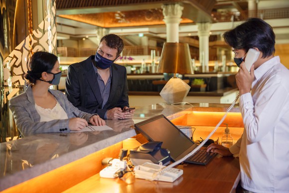 Two people, wearing masks, checking into a hotel while being helped by the hotel clerk, wearing a mask. 