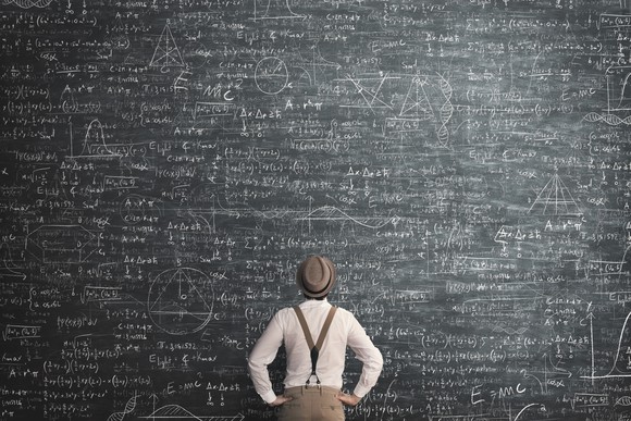 A man wearing suspenders standing in front of a giant chalkboard with equations.
