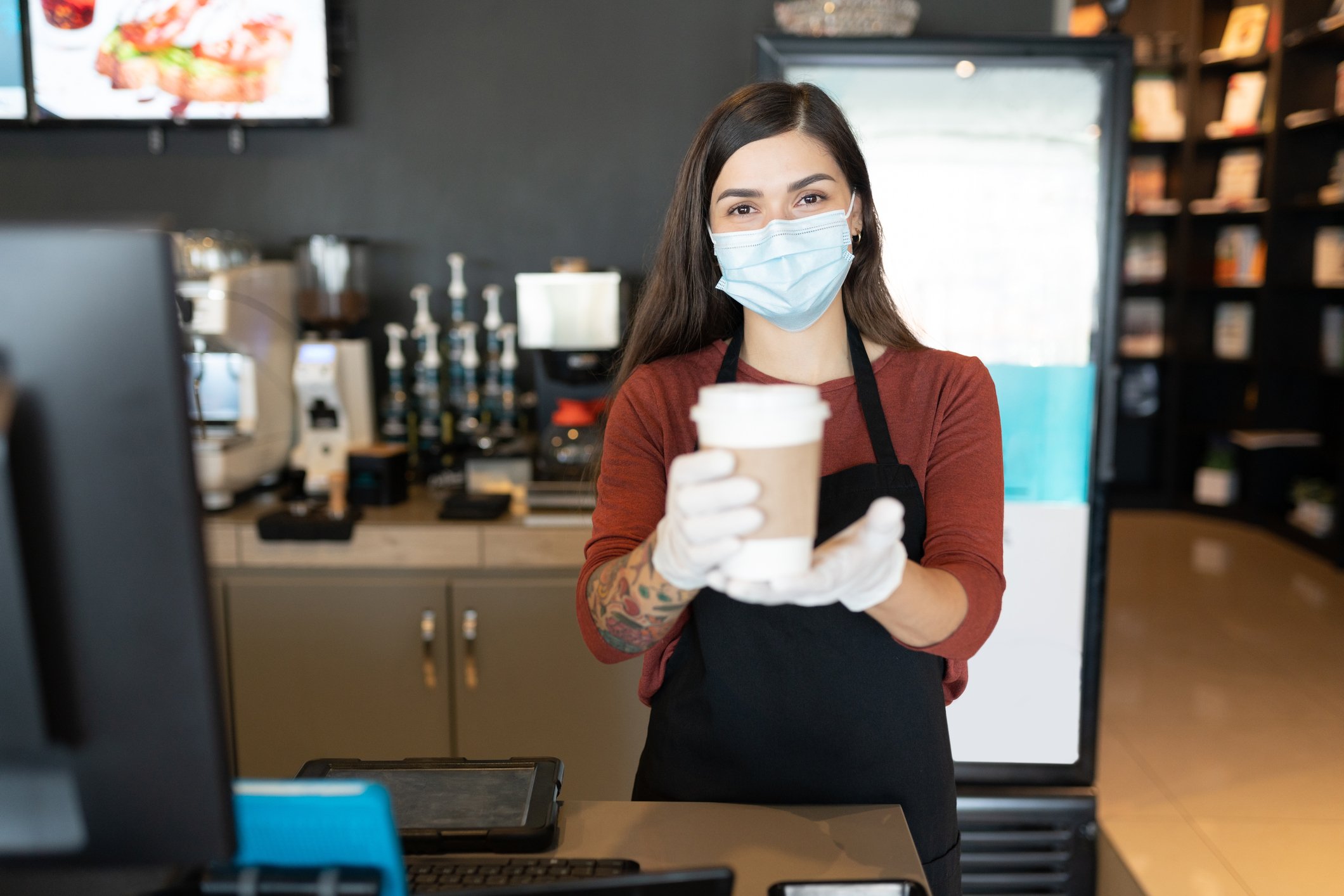 Barista wearing face mask, serving a beverage in a paper cup.