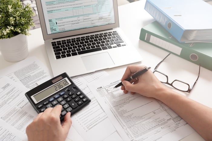 Person filling out form next to laptop while typing on calculator