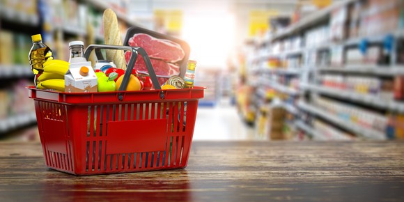 A red plastic shopping basket full of supermarket food items, resting on a wood surface with grocery shelves in the background.