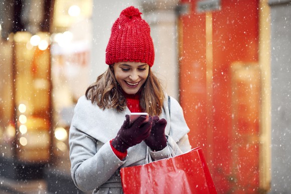 Smiling woman with shopping bags looking at phone in snow