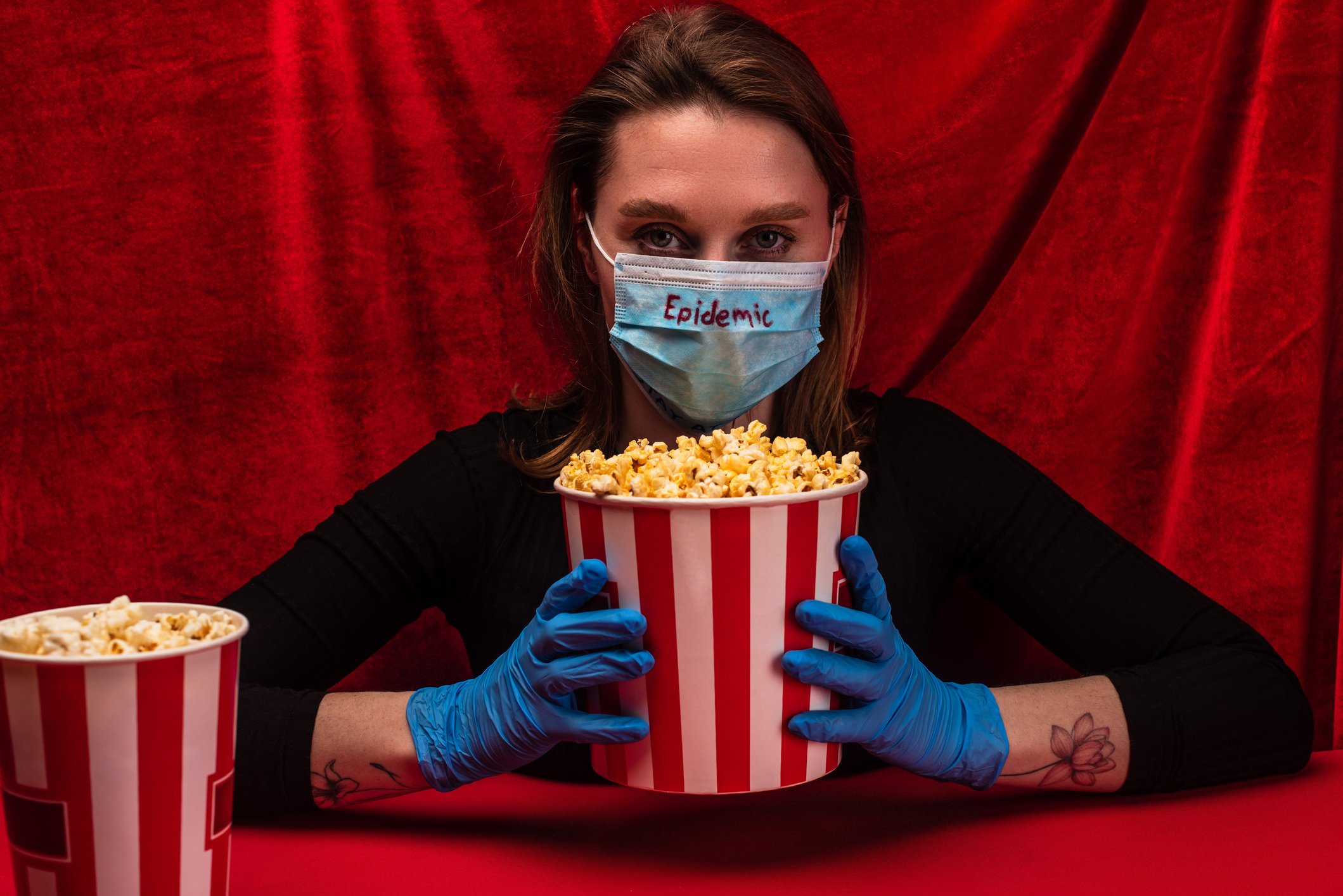 A woman in a face mask and latex gloves selling movie theater popcorn against a background of red drapes.