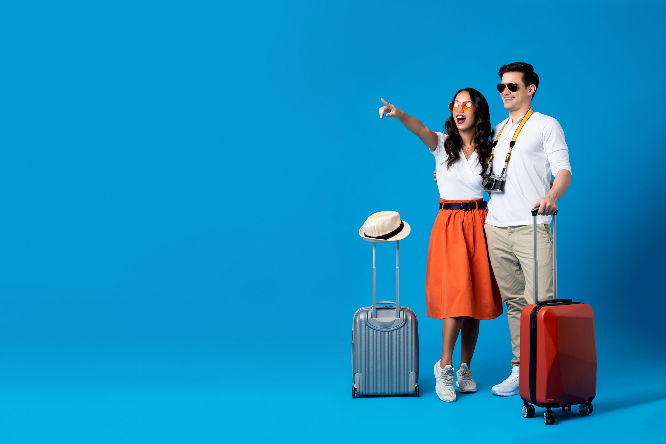 A young couple with suitcases against a blue background. The woman points to something in the distance. 