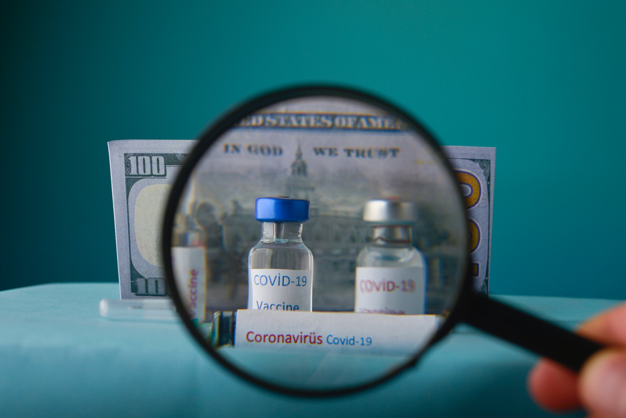A magnifying glass held in front of two COVID-19 vaccine vials with a $100 bill behind them