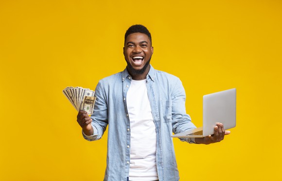 A smiling man holding cash in one hand and a laptop computer in the other