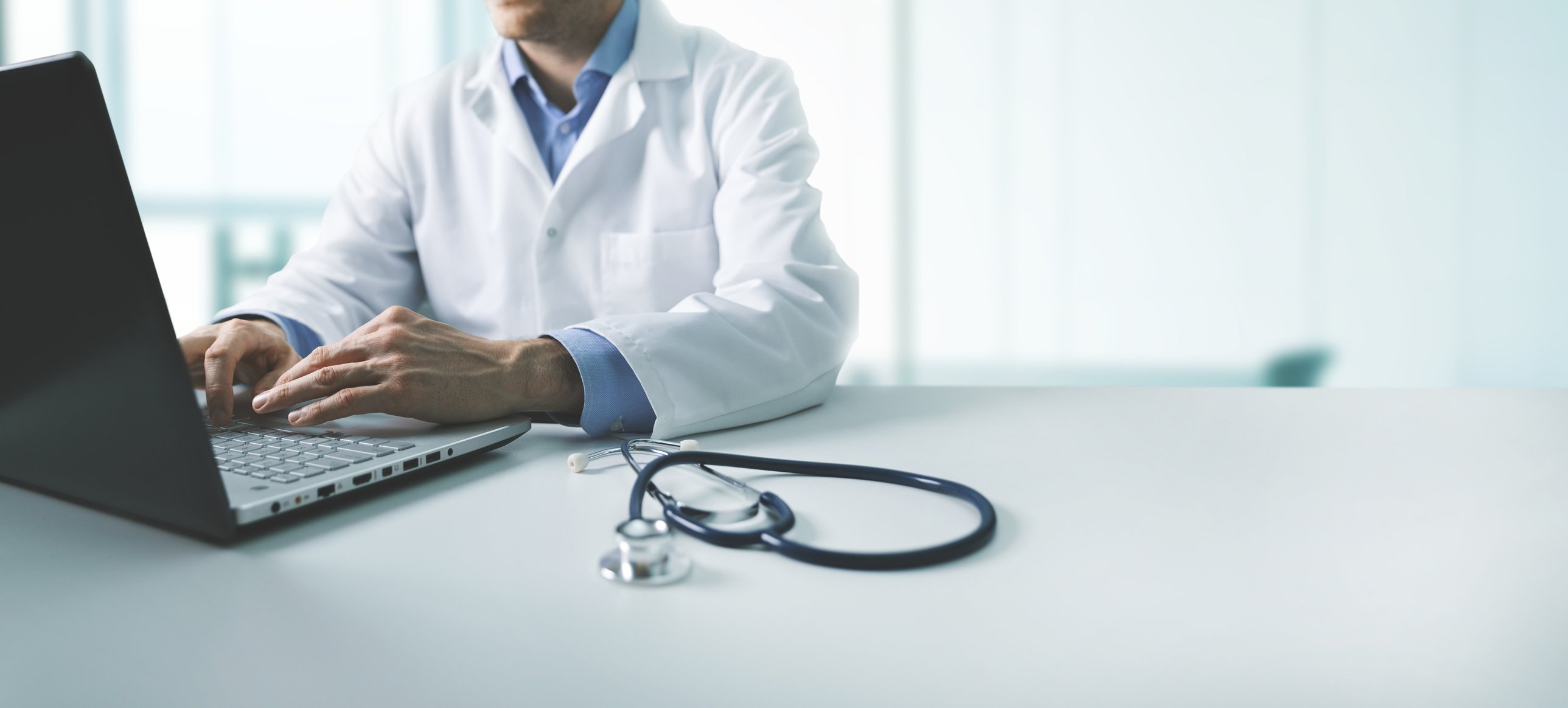 Doctor in lab coat using a laptop, with stethoscope in the foreground on the table.