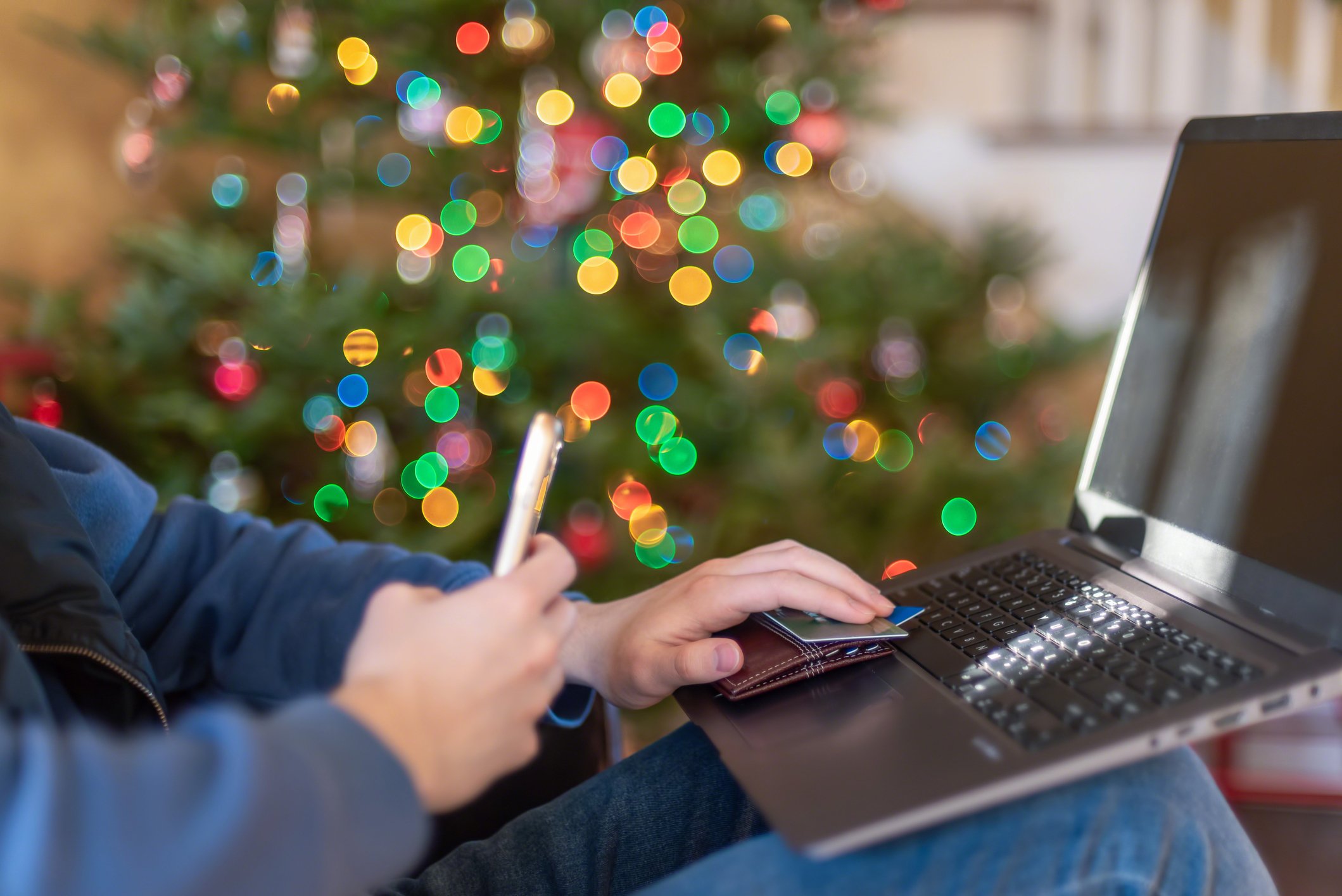 A man at a computer in front of a Christmas tree with his wallet and credit card out, making an online purchase. 