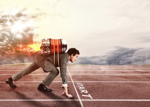 A businessman crouches at the starting line like a sprinter, wearing a fired-up jetpack on his back.