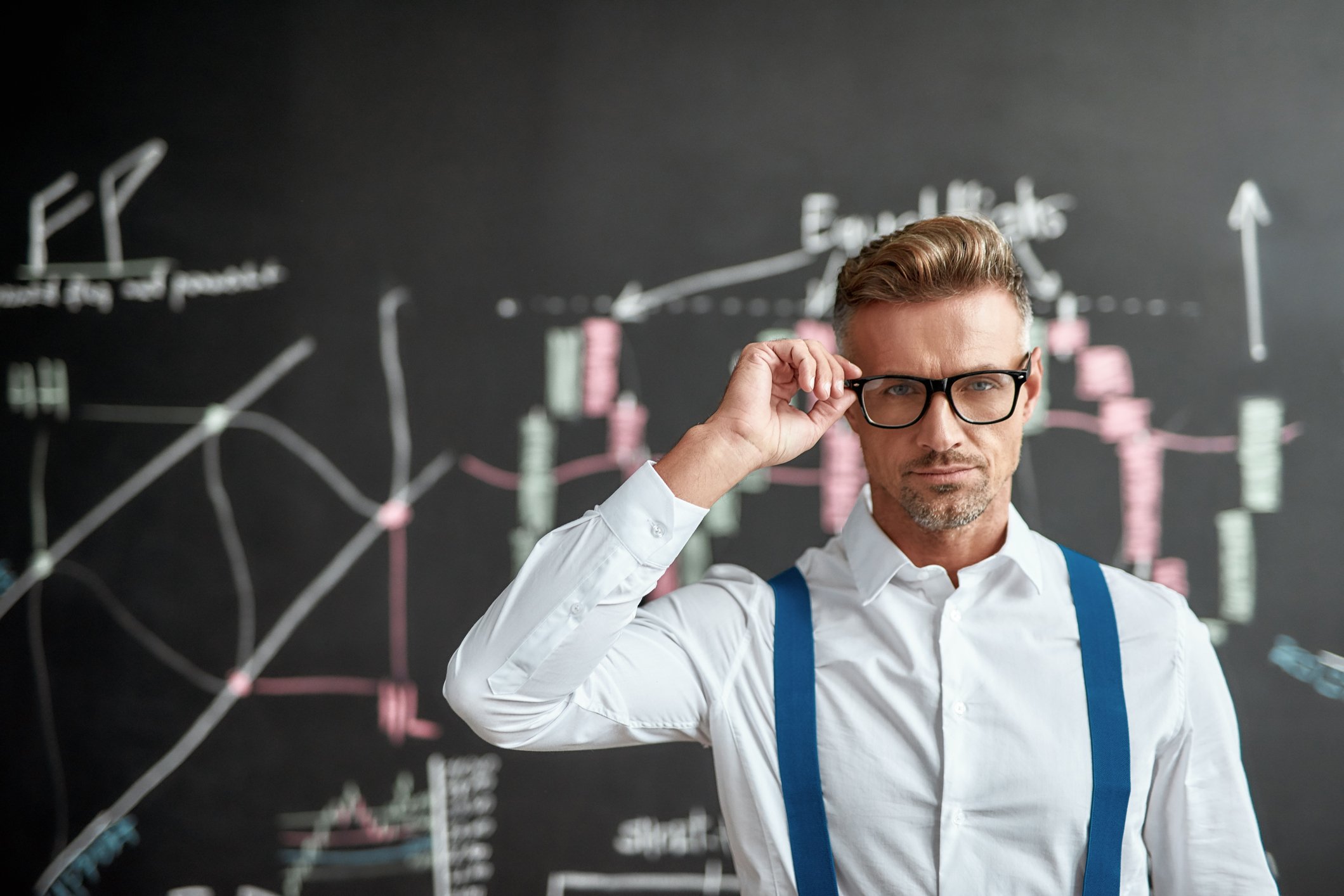 A trader wearing blue suspenders stands in front of a blackboard.