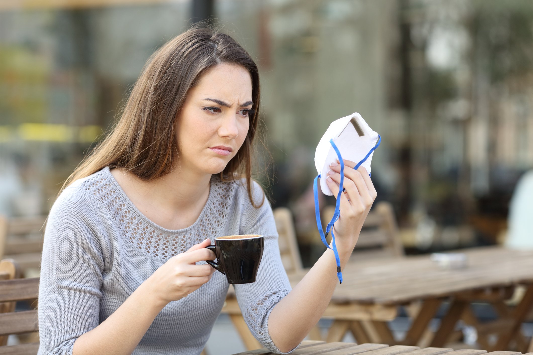A young woman frowns at her face mask, holding a cup of coffee in her other hand.