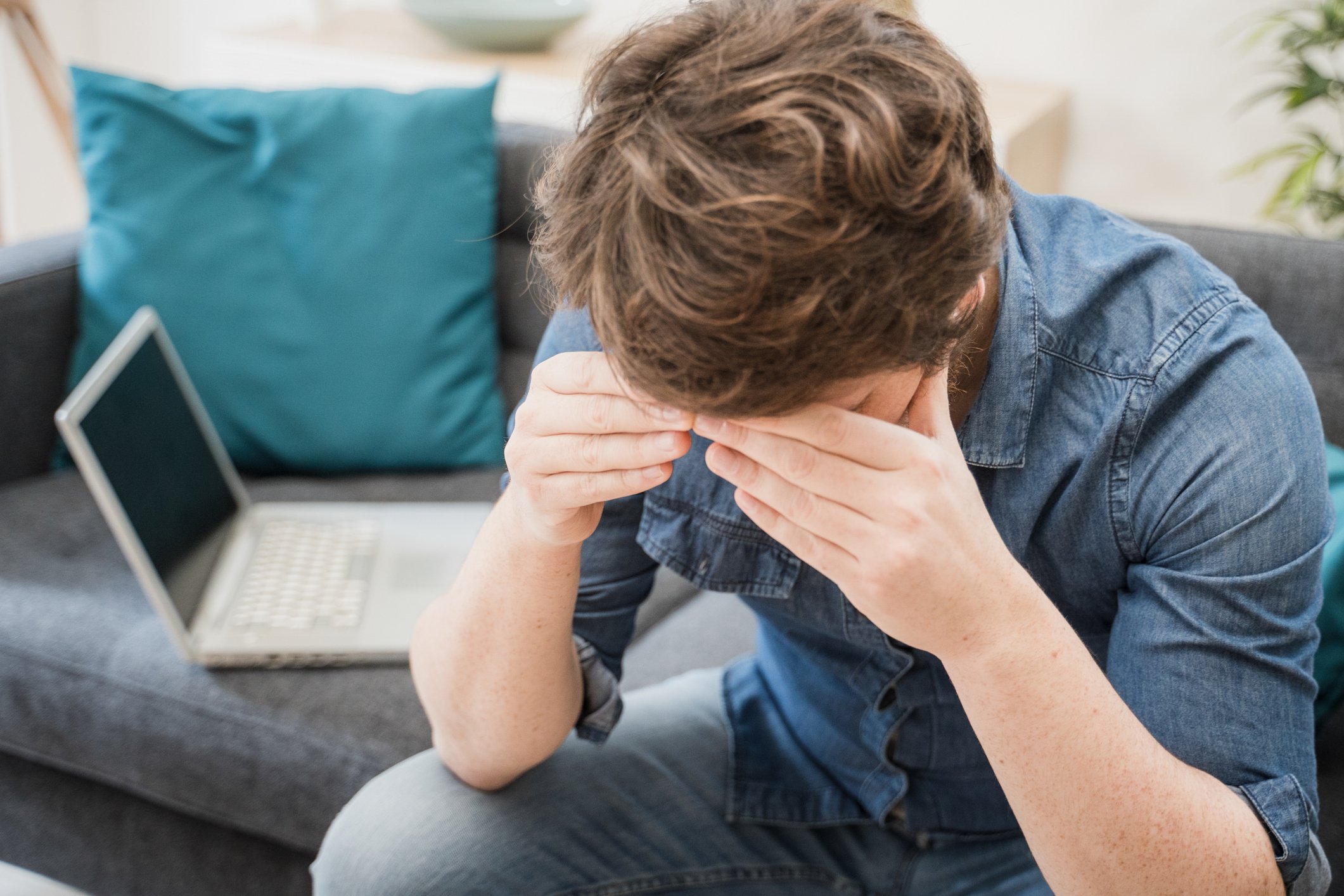 A disappointed investor holds his head as he sits on his couch with his laptop in the background.