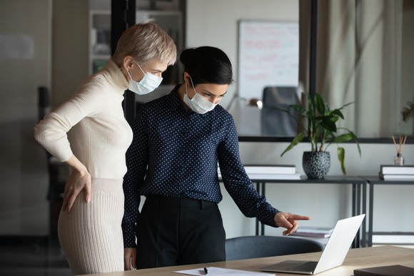 Two female medical executives wearing protective masks look at a laptop computer in a modern office.