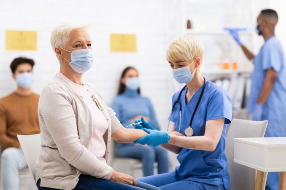 A masked nurse administering a vaccine to a masked elderly woman in a room with masked people waiting.