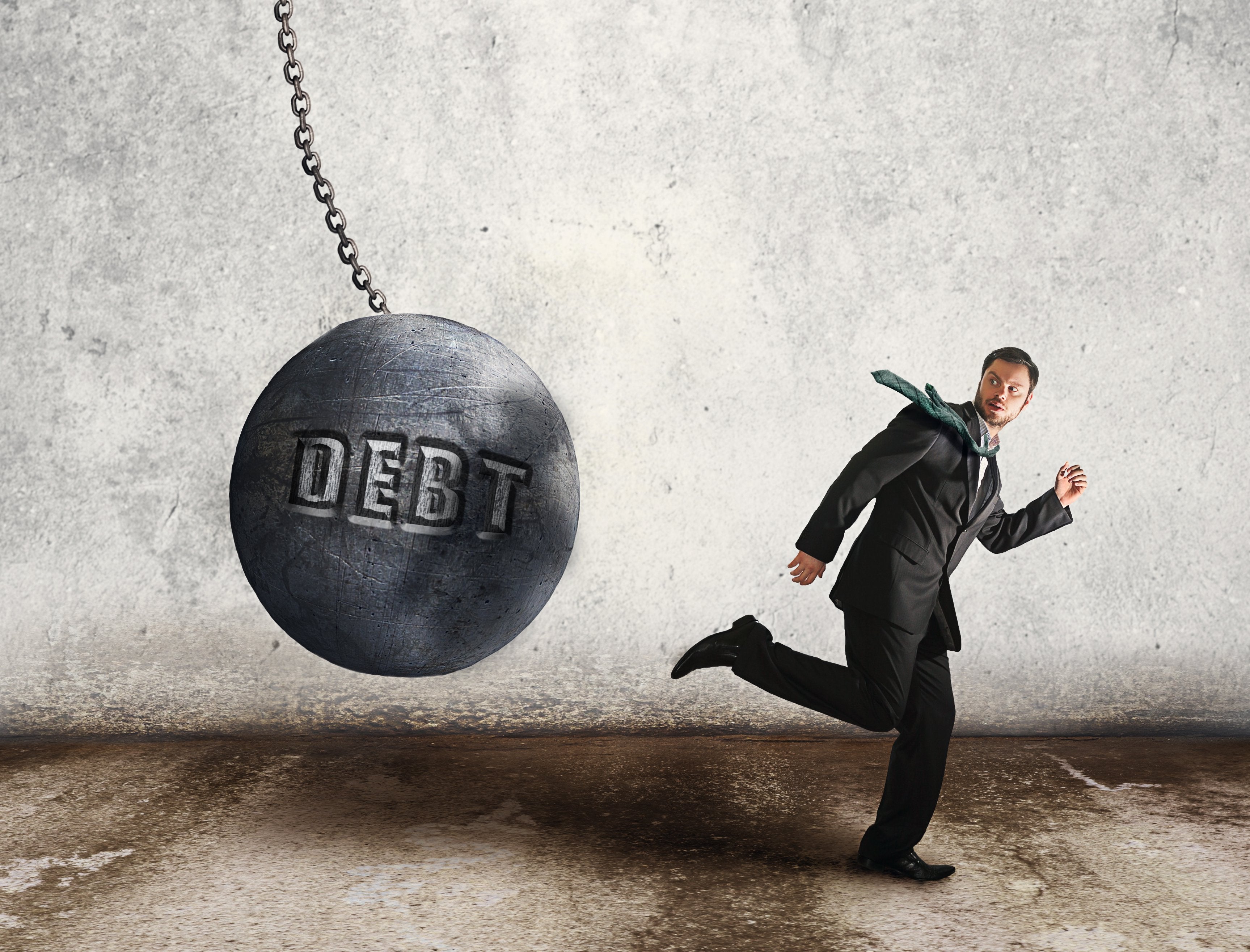 The word Debt on a wrecking ball swinging toward a man running away.