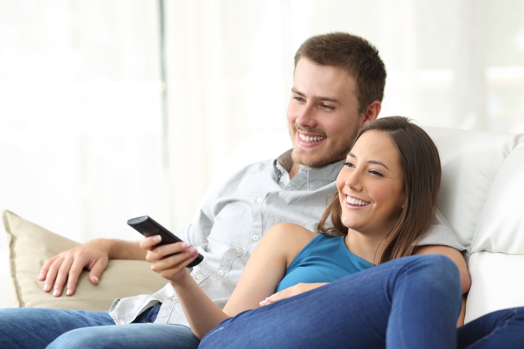 A smiling young couple cuddle up on the couch with a TV remote.