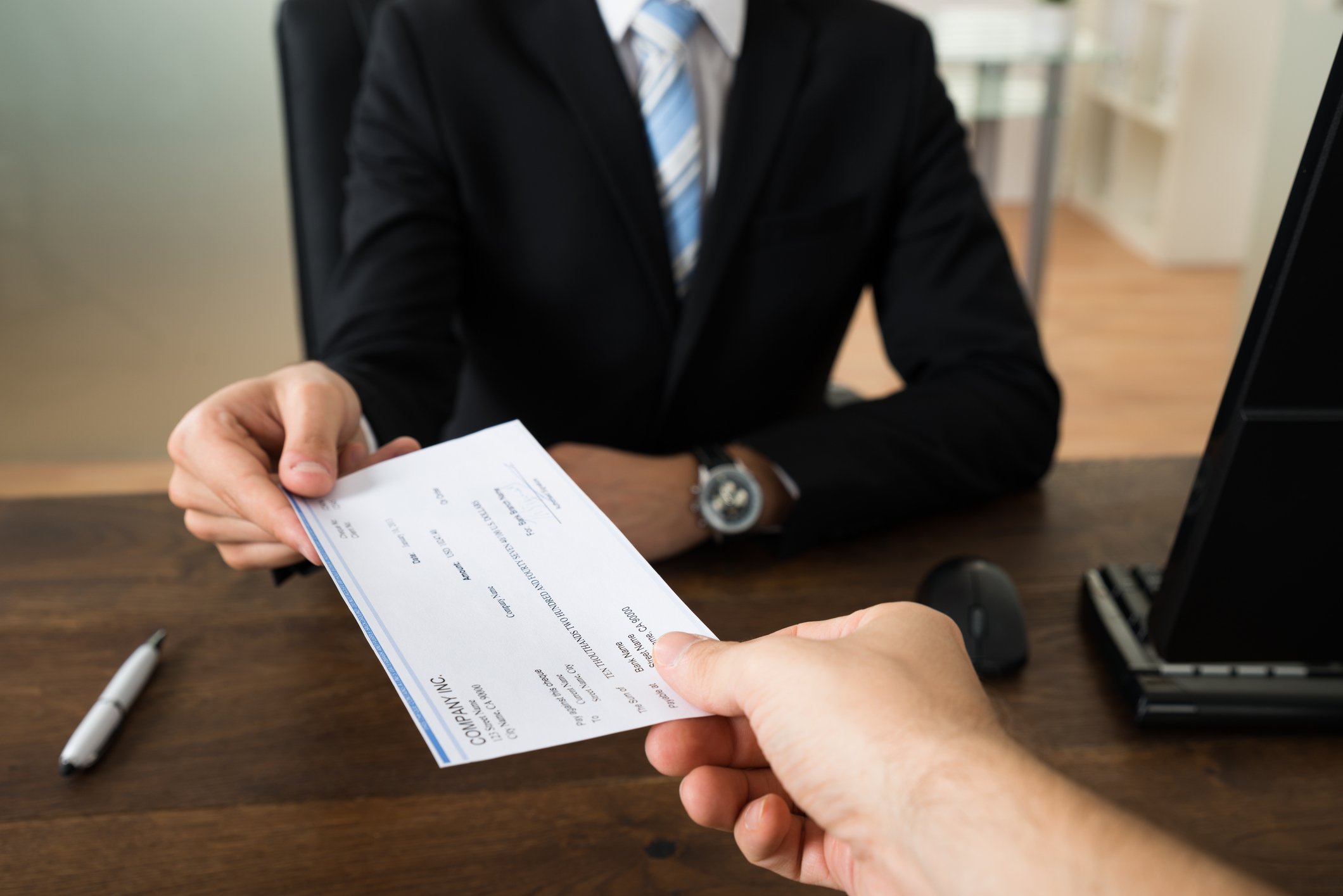 Businessman seated at a desk handing over a check to an unseen peer.