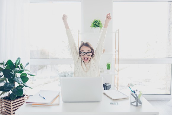 A woman celebrating with hands in the air from her desk.